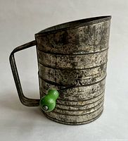 Side view of the tin flour sifter showing metal ribbed body and green knob on hand crank.