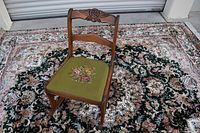 Full view of the antique wooden rocking side chair with floral needlepoint seat, placed on a patterned carpet outdoors.