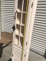 Full side angle view of the white corner curio cabinet showing glass door with inner shelves and black tassel handle, tape measure vertically to indicate height dimension.