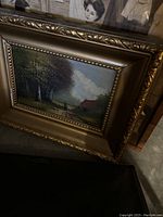 Framed oil painting showing a rural scene with trees, a figure on a path, and a small house under a cloudy sky in a decorative gold frame.