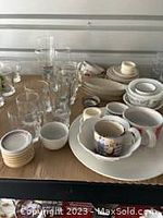 Photo showing the entire lot of assorted glassware and pottery on a cardboard surface, including clear glasses, pottery cups with saucers, and small dishes.