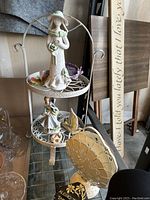 Photo of a two-tier white metal stand holding various small statues including a woman figurine, children figurines and a butterfly, along with a heart-shaped napkin holder in the front.