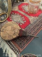 Photo showing a corner of the red Persian rug with geometric central medallion, fringed edges, and part of a vintage fireplace bellow lying on it. The bellow has detailed artwork of children on leather facing and a metal nozzle.