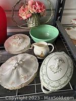 Multiple vintage porcelain serving pieces on metal shelving including intricate covered dishes, pitcher with figure design, and green bowl.