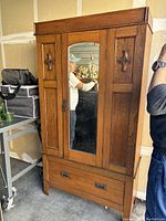 Full view of solid oak cabinet showing front with mirrored central door and decorative side panels, standing on legs, drawer at bottom.
