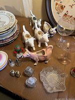 Photo shows porcelain cow pitchers, assorted salt and pepper shakers including novelty designs, depression glass goblets, clear patterned glass bowl, and stacked vintage plates with floral and animal motifs