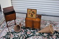 Photo showing vintage wooden standing magazine rack, wooden two-door storage cabinet, framed floral picture, and two large woven cornucopia baskets on floral rug.