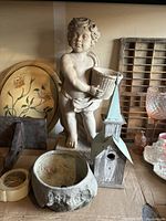Photo of cherubic child statue holding basket, vintage birdhouse, and clay bowl on shelf with other items.