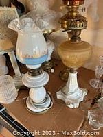 Two vintage table lamps on display; one white porcelain and milk glass with blue and gold floral glass shade, the other beige milk glass with gold details and brass fittings.