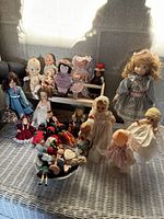Photo showing a variety of vintage dolls in different sizes and detailed outfits, arranged on a wicker surface with natural sunlight casting shadows.
