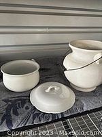 Two white pottery items: large pot with wire handle and wooden grip, and smaller covered bowl with lid