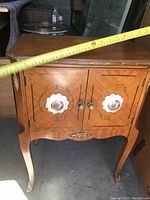 Front angle view of the vintage side table showing the wooden inlay door fronts, painted porcelain knobs, and curved legs.