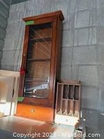 Photo showing full wooden storage cabinet with glass front and bottom drawer, wood warm toned and varnished.