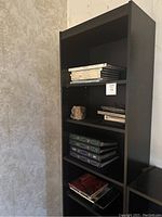 Front view of the black wooden bookcase against a light-colored wall, displaying the top three shelves with books and a decorative stone.