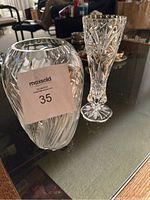 Photo showing two crystal vases on a glass tabletop, one large with swirling wave design and one smaller with pinwheel cut design.