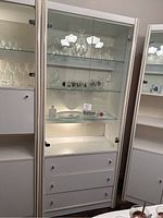 Photo of two white dining room cabinets featuring glass doors with shelving inside, interior lighting, and drawers below.
