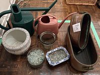 Shows two vintage oval copper herb planters with patina, a pink plastic watering can, a dark green watering can, white porcelain planter, glass jar, small blue and white rectangular porcelain pot, and glass bowl with marbles.