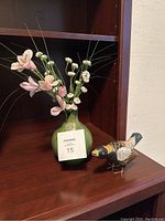 Green ceramic vase with artificial pink and white flowers and long greenery strands, next to a bird figurine, all set on a wooden shelf.