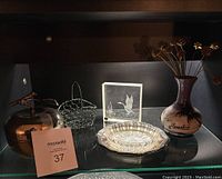 Display of miscellaneous decorative pieces on glass shelf including a glass art block, metal basket, ceramic vase, silver tray, and metallic cloche.