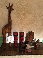 Front view showing all items on a wooden plank including giraffe, two red head carvings, natural wood sculpture, bird, small animal figurine, and container.