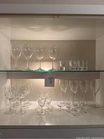 Shelves with clear glassware including 8 tall wine glasses, 5 smaller wine or cordial glasses, and 5 shot glasses all in very good condition.