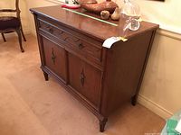 Front and side view of wooden cabinet/server showing ornate metal pulls, one drawer, and two doors with decorative panels.