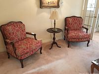 Two matching bergère style armchairs with carved dark wood frames and red floral damask upholstery flanking a small round side table with lamp
