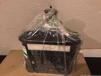 Front view of the black mailbox wrapped in clear plastic, showing the peaked top and label holder.