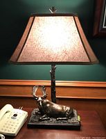 Table lamp illuminated on, showing bronze-colored sculptural base with reclining moose and fabric shade emitting warm light.