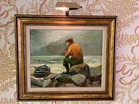 Full view of the framed oil painting showing a man sitting on rocks by the sea next to a small boat, with a pipe in his mouth and the mounted lamp above the frame.