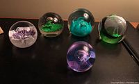 Five art glass paperweights on display showing different designs and colors.