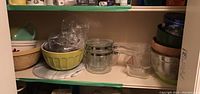 Full view of shelves showing yellow and beige ceramic mixing bowls, metal mixing bowls, double boiler glass pots with clear handles, glass loaf pan, and other kitchen items