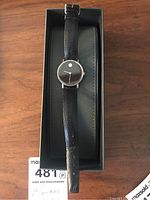 Photo of Movado watch inside original black box on wood surface, showing full watch and strap.