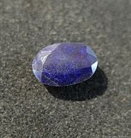 Close-up photo of front top facets of the blue sapphire gemstone on black surface showing oval shape and blue color.