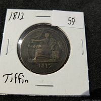 Front side of 1812 Tiffin token showing seated figure holding scales and dated 1812, secured in cardboard holder.