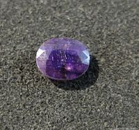 Close-up image of the oval-shaped faceted sapphire gemstone, showing its blue to purple gradient color and surface inclusions.