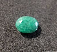 Close-up front angled view of green oval emerald gemstone showing faceted surface and natural inclusions on black textured background.