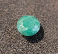Close-up photo of the loose emerald gemstone on a black textured surface, showing the stone's round cut and green color with visible inclusions.