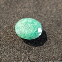 Close-up photo of the faceted emerald gemstone showing its vivid green color and surface faceting on a dark textured background.