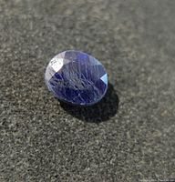 Close-up photo of oval blue sapphire with visible surface scratches and inclusions on textured background.