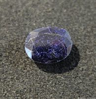 Front angled view of the oval blue sapphire gemstone on a dark textured background showing surface imperfections and inclusions.