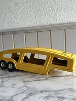 Photo of yellow 1960s Structo car carrier trailer on white marble surface showing side and wheel details.