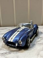 Front angled view of metallic blue 1964 Shelby Cobra 427S/C die-cast model with white racing stripes, showing grill, headlights, and windshield.