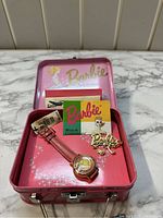 Pink metal Barbie-themed box containing a pink leather strap wristwatch, Barbie pin, and booklets.