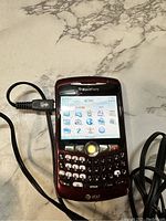 Front view of maroon Blackberry 8310 Curve showing screen with apps and charging cable plugged in.