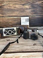 Olympus Camedia C-3000 Zoom digital camera with a zoom lens, black grip, buttons on top, and instruction manual in background. Next to it is a vintage 3-lens camera in a brown leather case.