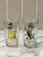 Two tall clear shot glasses on marble surface both featuring colorful Disney character prints: Snow White with Dopey on one glass, Jiminy Cricket on the other.