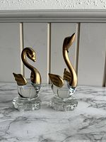 Photo showing two clear glass swan figurines with gold painted necks, heads, wings, and tails sitting on clear glass bases. The swan on the left has a curved neck, the one on the right has its neck upright.