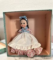 Front view of Madam Alexander Argentina doll wearing a floral dress with white apron and blue bow in hair, inside original box.