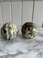 Two round porcelain decorative balls featuring marbled green, cream, black, and brown swirls with a glossy finish placed on a marble surface background.